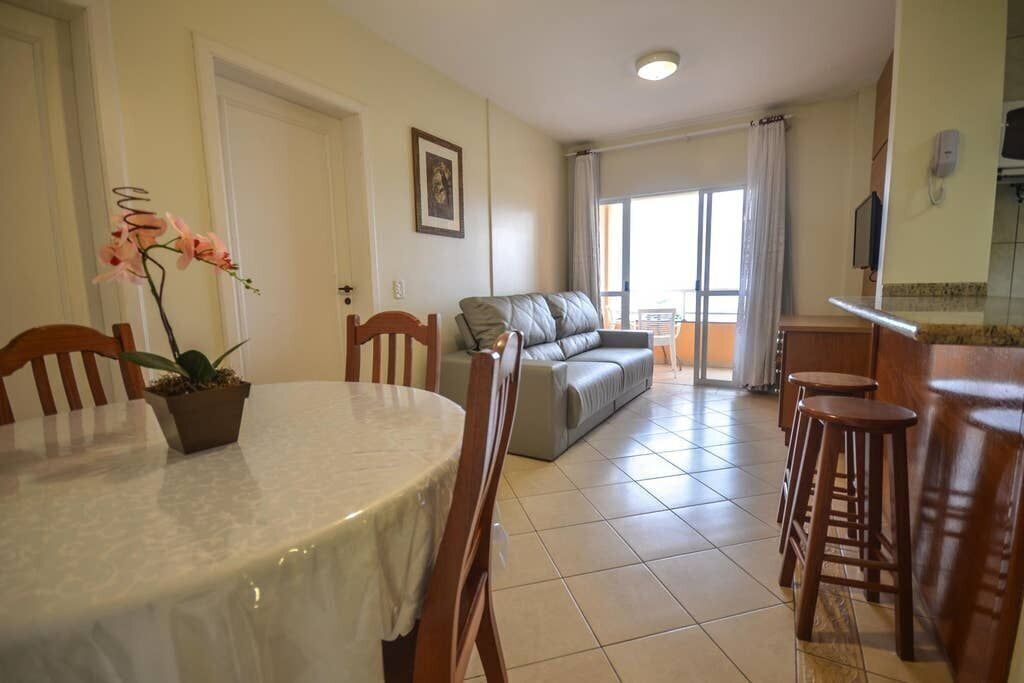 A dining area with a round table, chairs, and a potted plant. There is a sofa and a balcony with a view.
