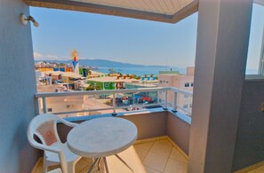 A balcony with a table and chairs, offering a view of a coastal town with colorful buildings and a lighthouse. - Residencial Vista Bella (Bombinhas)