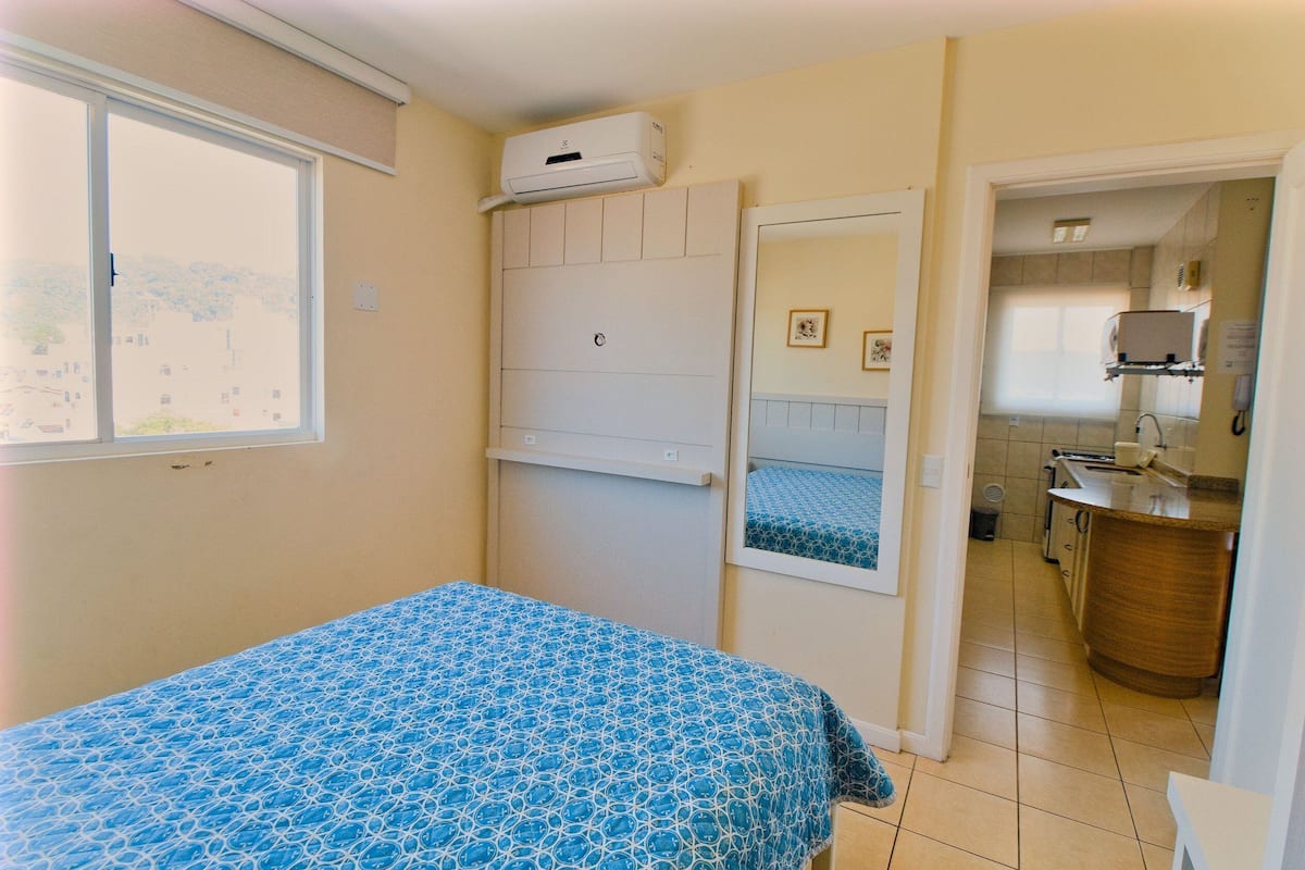 A bedroom with a bed, a window, a door leading to a kitchen, and an air conditioning unit.
