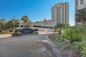A gated entrance to Beach Colony Resort with a sign, palm trees, and a parking garage in the background.