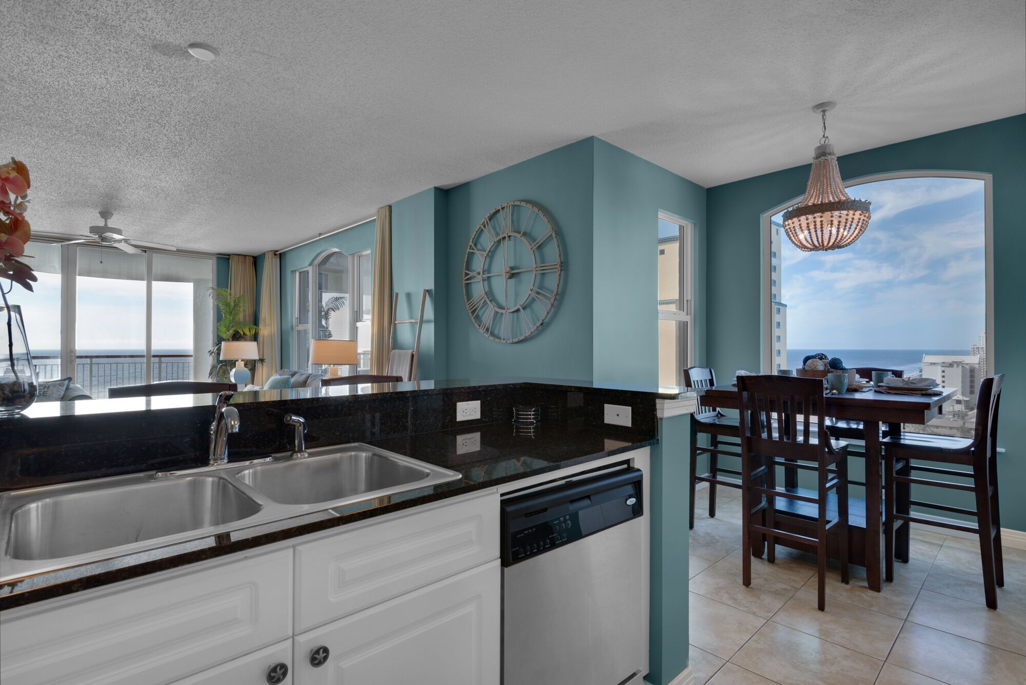 A modern kitchen with a black countertop, stainless steel sink, and a dishwasher. There is a dining area with wooden chairs and a table, and a large window offering a view of the ocean.