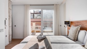 Ein Hotelzimmer mit einem Bett, einem hölzernen Kopfende, einem Nachttisch, einer Wandlampe und einem Balkon mit Blick auf Gebäude.