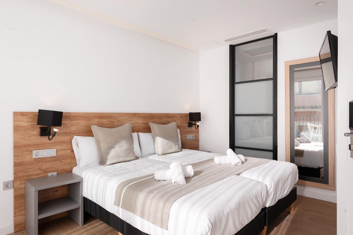 Une chambre d’hôtel moderne dotée d’un grand lit, d’une tête de lit en bois, de deux tables de chevet, d’un téléviseur fixé au mur et d’une porte-fenêtre coulissante donnant sur un balcon.