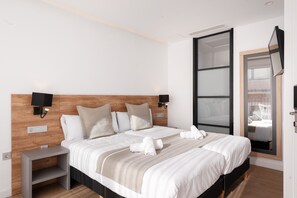 Ein modernes Hotelzimmer mit einem großen Bett, einem Holzkopfende, zwei Nachttischen, einem an der Wand montierten Fernseher und einer Glastür zum Balkon.