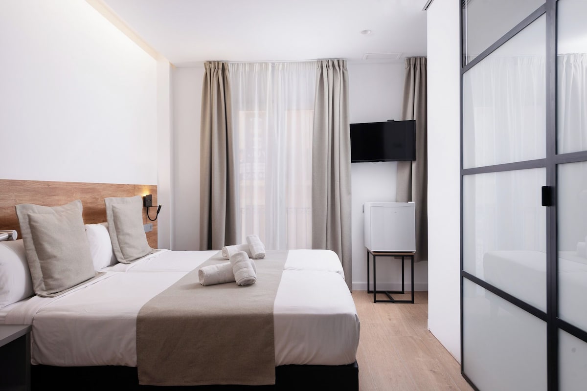 Une chambre d’hôtel avec un lit, deux oreillers, une table de chevet, une télévision et une porte-fenêtre coulissante.