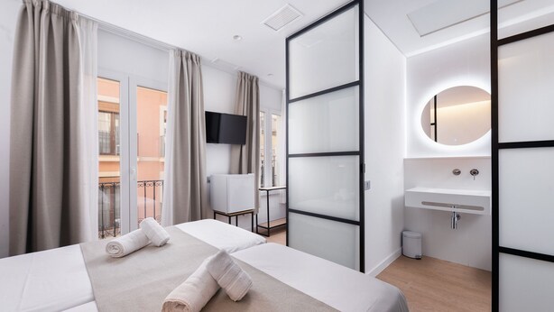 Ein Hotelzimmer mit Bett, Fernseher, Bad mit Waschbecken und Spiegel sowie Balkon mit Vorhängen.