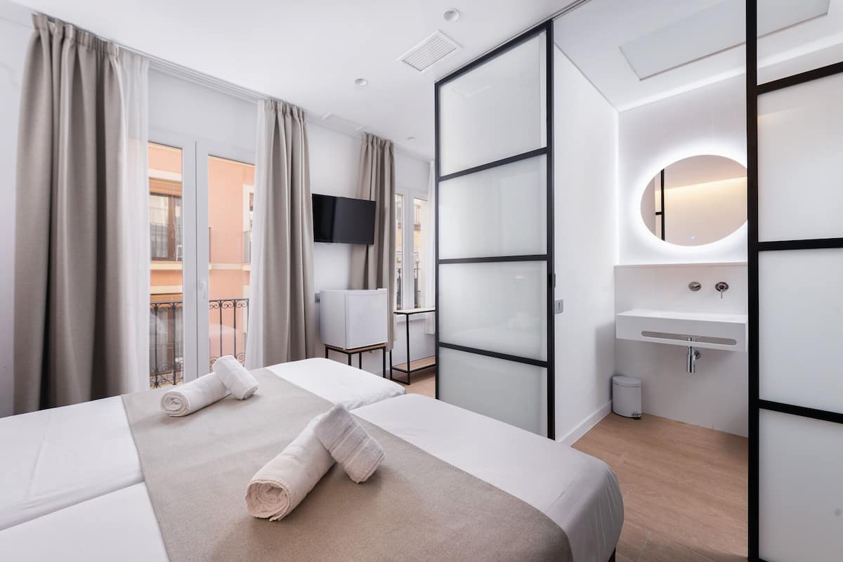Une chambre d’hôtel moderne équipée d’un lit, d’une télévision, d’une salle de bain avec un lavabo et un miroir, et d’un balcon avec des rideaux.