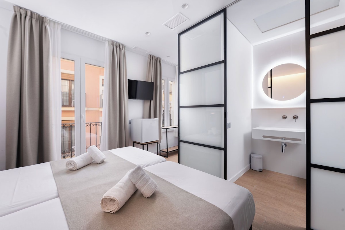 A hotel room with a bed, a TV, a bathroom with a sink and mirror, and a balcony with curtains.