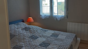 A bedroom with a large bed, a window with curtains, a bedside lamp, and a radiator. - single-storey house 600 m beach (Étables-sur-Mer)