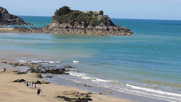 A coastal scene with a rocky island featuring a building, clear blue water, and a sandy beach with people. - single-storey house 600 m beach (Étables-sur-Mer)