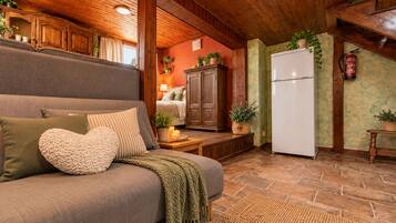 A cozy living room with a sofa, a fireplace, a kitchen area with a refrigerator, and wooden beams on the ceiling.
