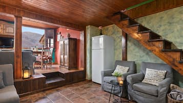 Een gezellige woonkamer met een houten plafond, een trap, een grijze bank, een kleine tafel met een kaars en een koelkast.