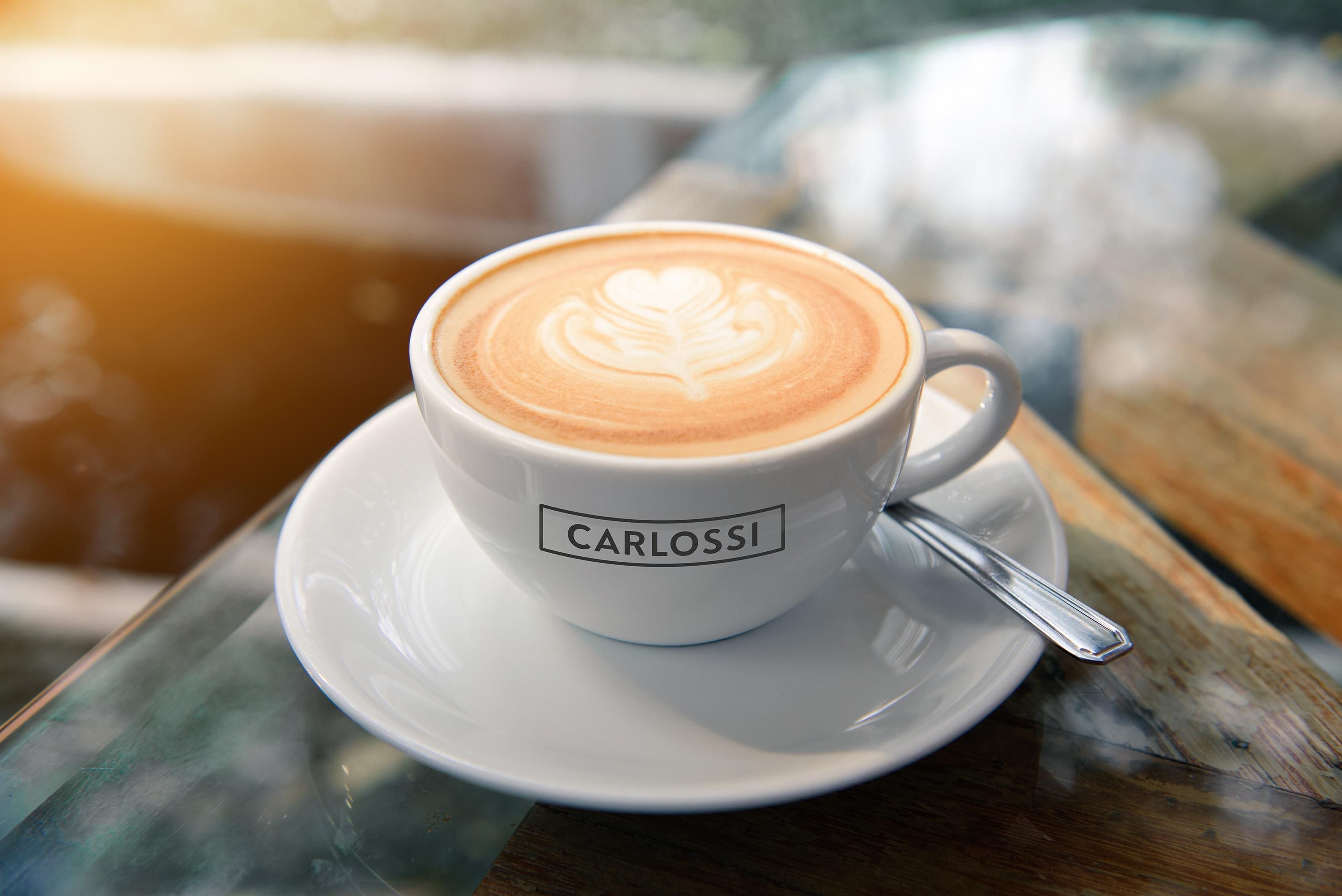 A white cup with latte art and the name 'CARLOS' on a saucer, placed on a wooden table.