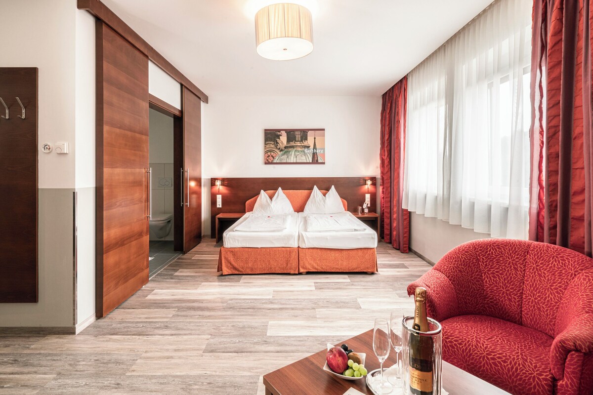 Habitación de hotel con cama doble, un sofá rojo, una mesa de centro de madera con una botella de champán y un cuadro en la pared.