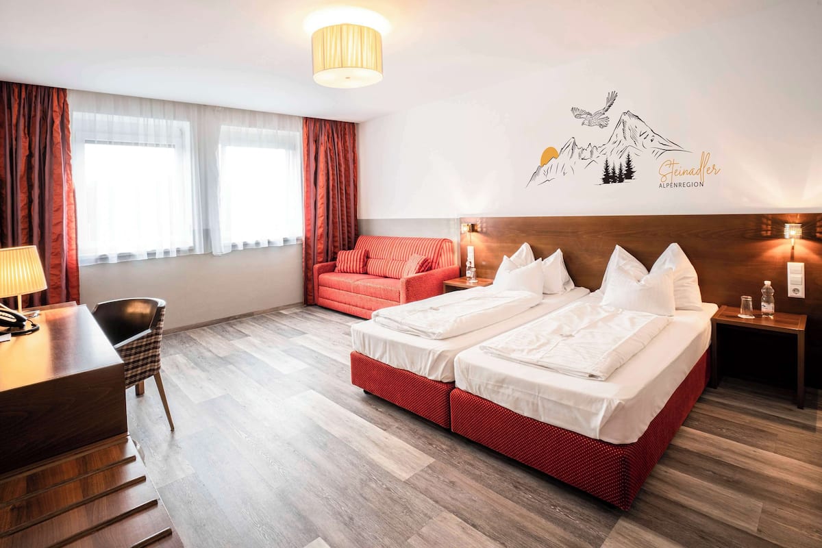Habitación de hotel con suelo de madera, un sofá rojo, dos camas con ropa blanca, un escritorio con lámpara, y un mural en la pared que muestra montañas y un pájaro.