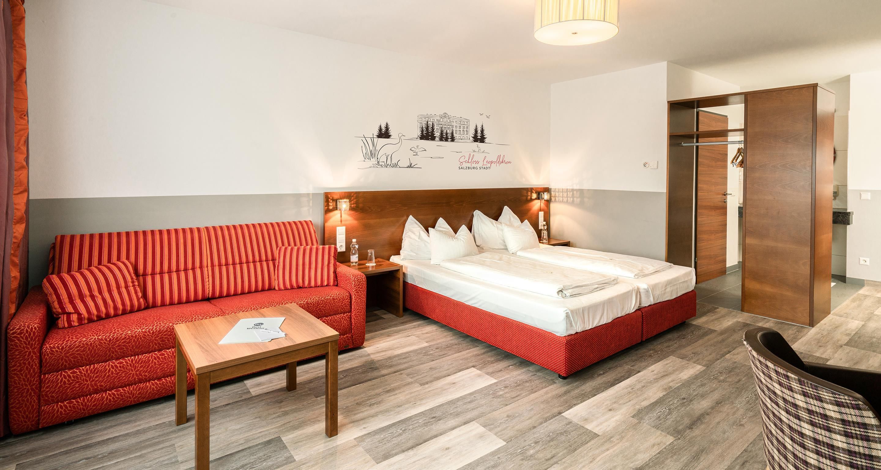 Quarto de hotel com sofá vermelho, mesa de madeira, cama com roupa de cama branca e uma obra de arte na parede.