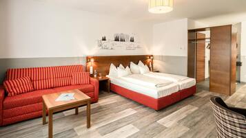 Quarto de hotel com sofá vermelho, mesa de madeira, cama com roupa de cama branca e uma obra de arte na parede.
