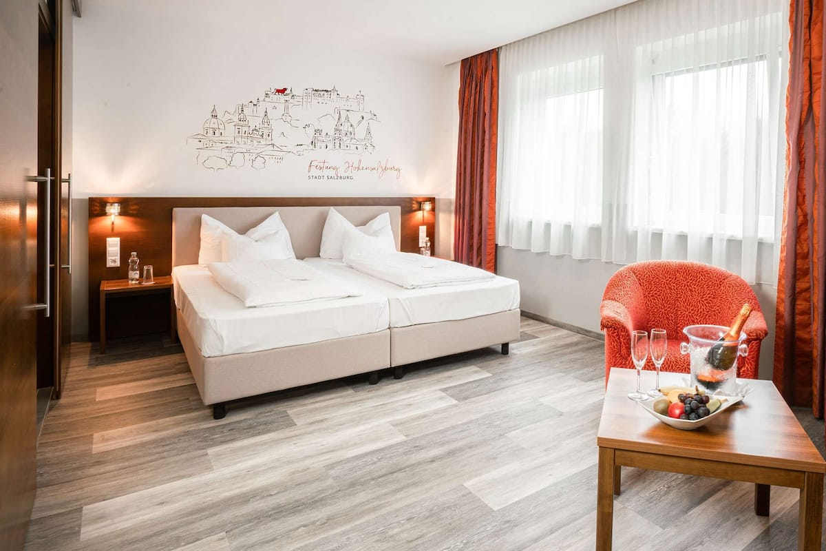 Una habitación de hotel con una cama, un sillón rojo, una mesa con fruteros y champán, y un mapa mural.