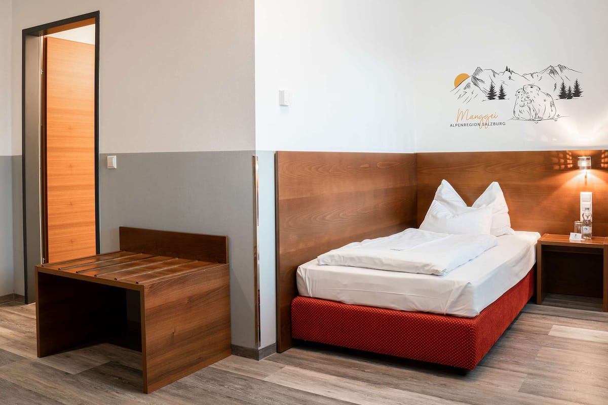 Una habitación de hotel con una cama de madera, un pequeño banco de madera, una mesita de noche con lámpara y un adorno en la pared.