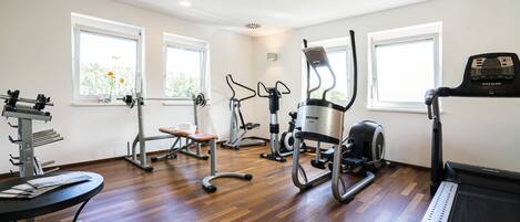 Un gimnasio con diversos equipos de ejercicio, amplias ventanas y suelo de madera.