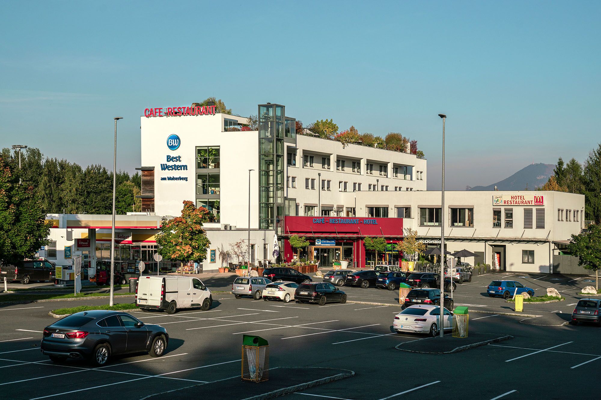 Un edificio de varios pisos con una cafetería, restaurante y hotel en la planta baja. Se ven varios coches aparcados delante y una gasolinera a la izquierda.