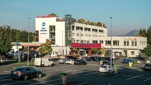 Un edificio de varios pisos con una cafetería, restaurante y hotel en la planta baja. Se ven varios coches aparcados delante y una gasolinera a la izquierda.
