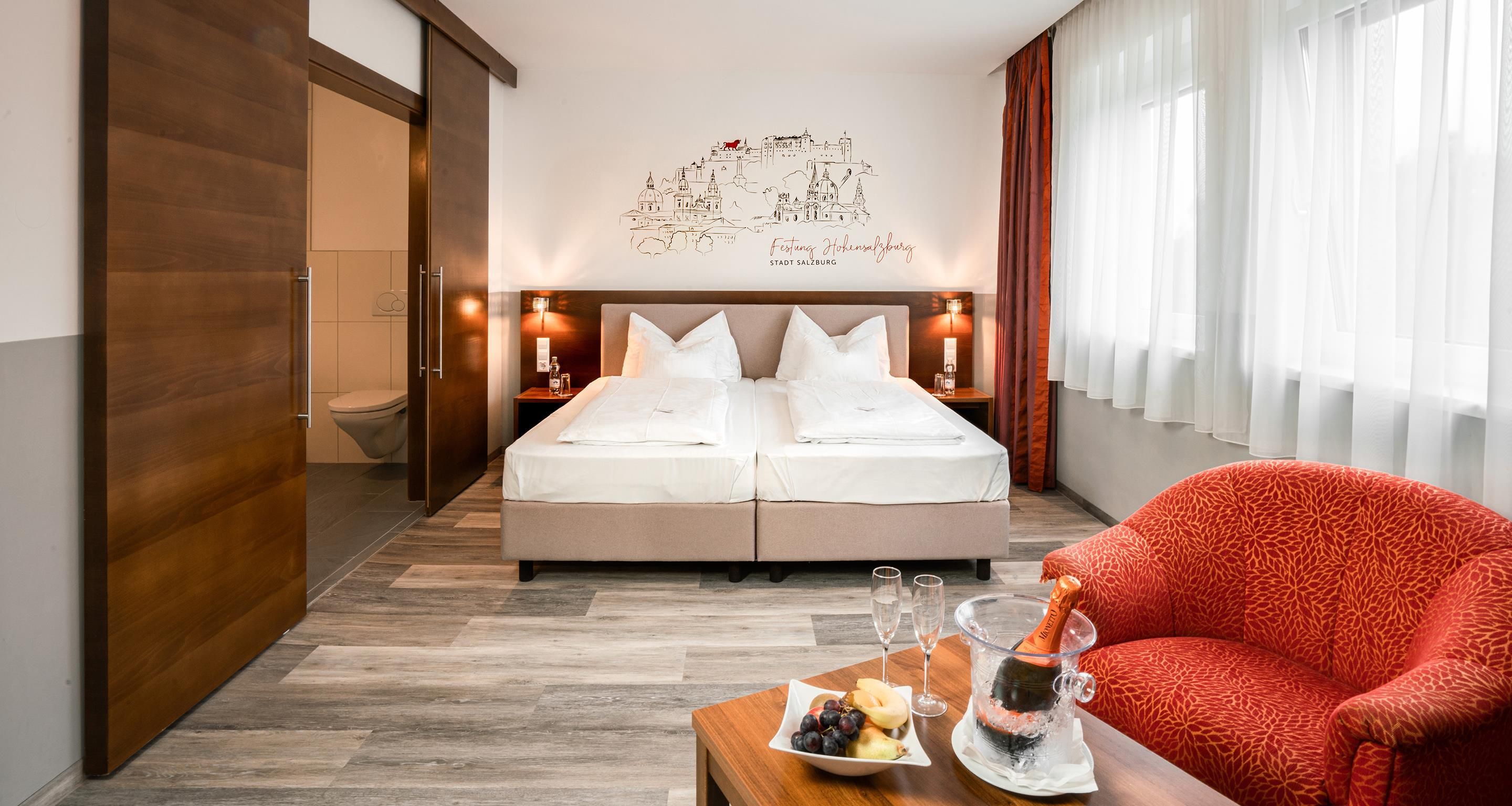 Hotellihuone, jossa on sänky, punainen nojatuoli, pöytä, jonka päällä on samppanjapullo, ja seinällä taideteos.