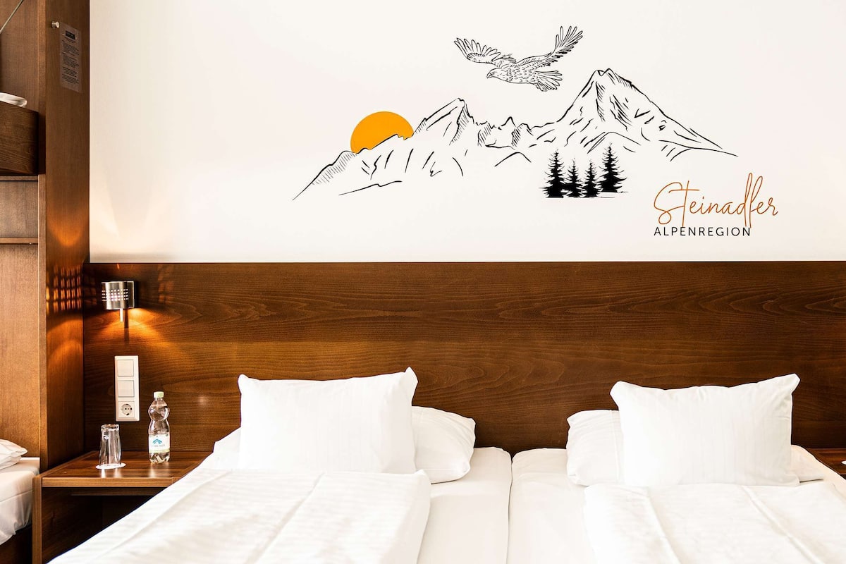 Habitación de hotel con cabecera de madera, dos camas blancas, un cuadro con montañas y un águila, y un cartel que dice 'Steinader Alpenregion'.