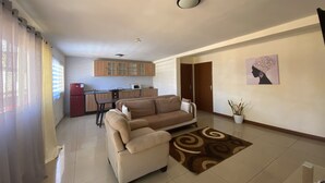 A living room with a beige sofa, a dining table with chairs, a kitchen area with cabinets, and a potted plant. - Residence Maeva (Mamoudzou)
