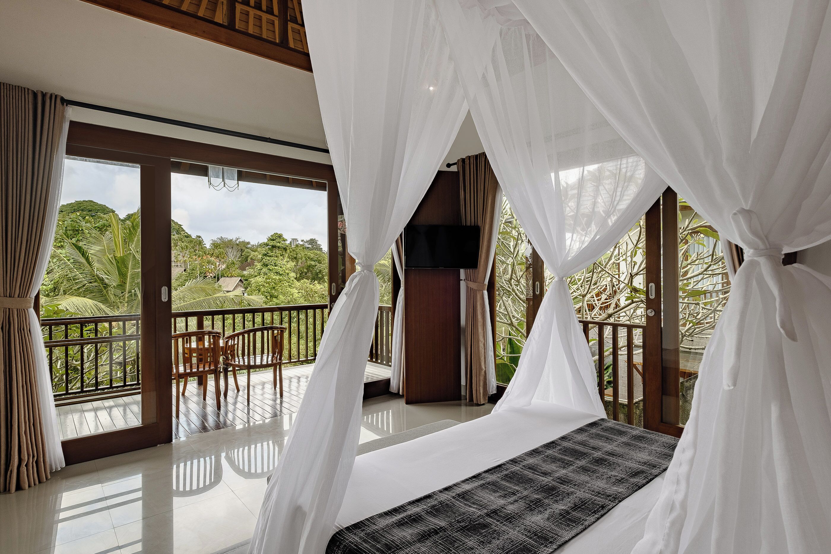 A room with a bed under a canopy, a TV, and a balcony with a view of greenery.