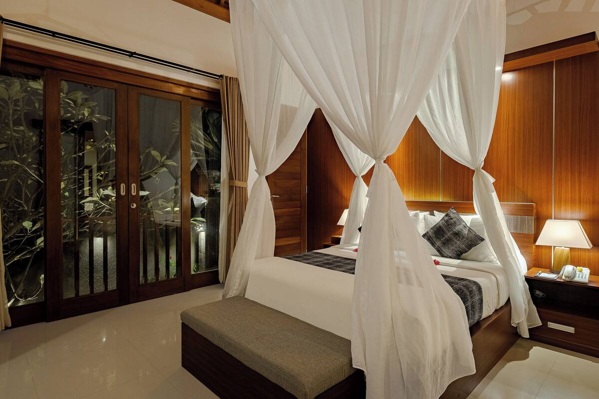 A four-poster canopy bed with a bench at the foot, a bedside table with a lamp, and a view of a balcony with plants.