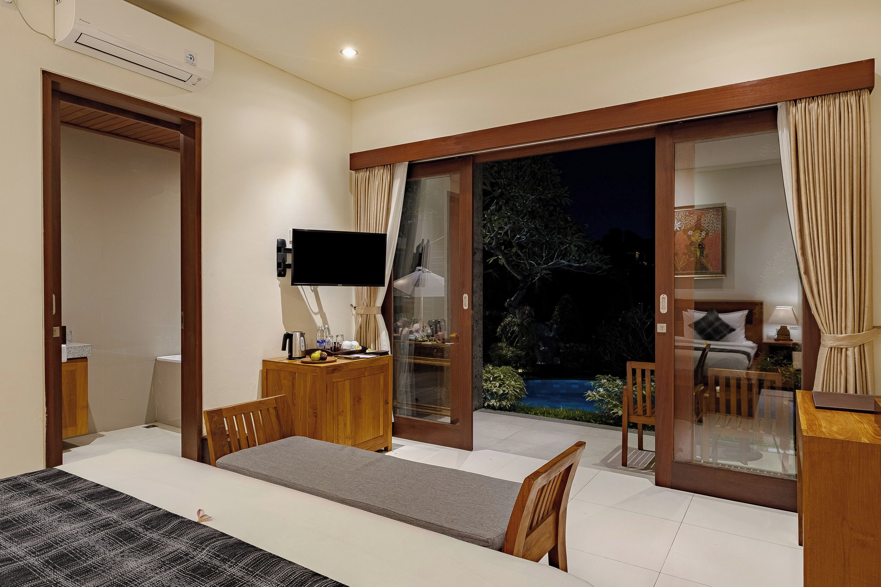 A bedroom with a bed, a desk, a TV, and a sliding glass door leading to a balcony.