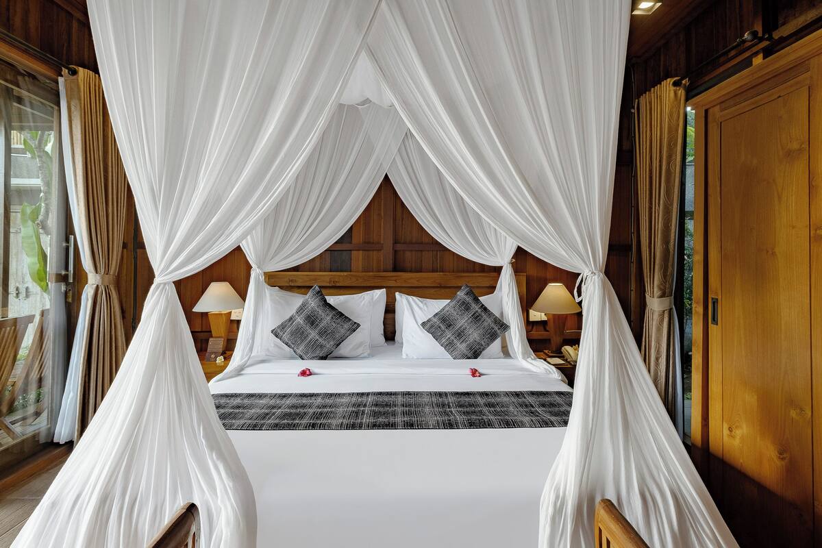 A four-poster bed with white drapes, two bedside lamps, and a wooden headboard.