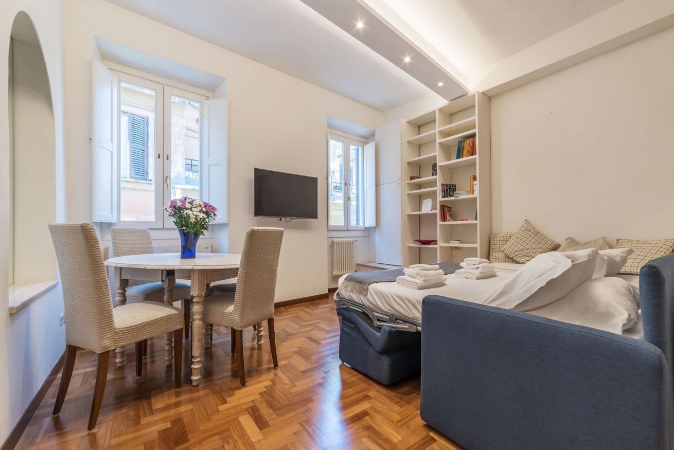 A modern room with a wooden floor, a round dining table with chairs, a bed with a blue headboard, and a bookshelf.