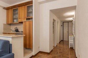 A compact kitchen with wooden cabinets and a built-in island, a hallway with a wooden floor, and a white door at the end.