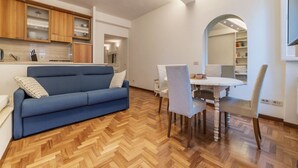 Ein modernes Wohnzimmer mit einem blauen Sofa, Holzparkettboden, einem Essbereich mit weißem Tisch und Stühlen sowie einer Küche mit Holzausstattung und einem Spiegel.
