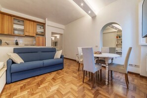 A modern living room with a blue sofa, wooden herringbone flooring, a dining area with a white table and chairs, and a kitchen with wooden cabinets and a mirror. - Lovely one bedroom apartment - Wifi, Independent Heating, A/C, Smart TV (Roma)