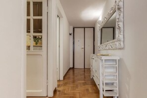 A hallway with a wooden floor, a white door, a white dresser with shelves, and a mirror with an ornate frame.