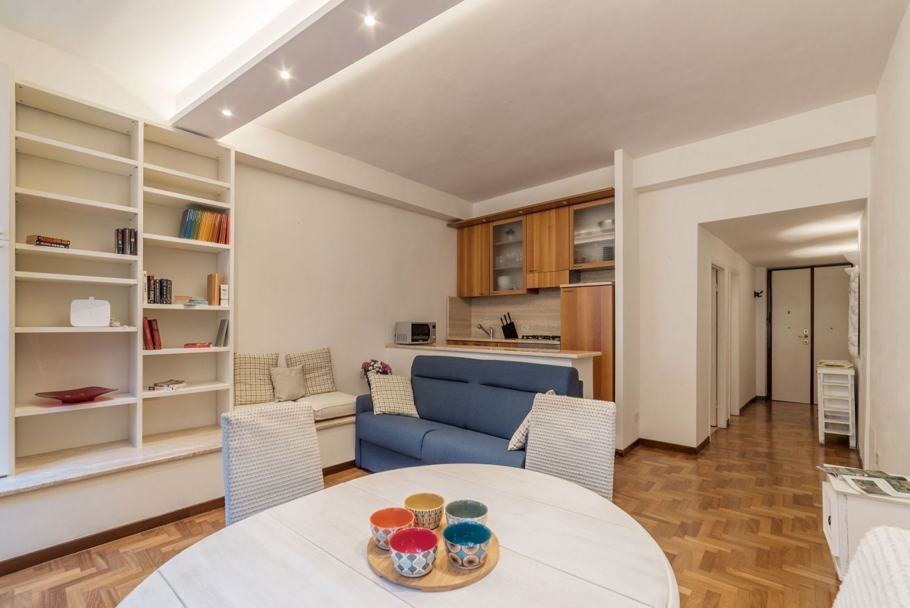 A modern living room with a blue sofa, a white coffee table with colorful bowls, a bookshelf, and a kitchen area with wooden cabinets.