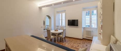 A modern living room with a dining area, a TV mounted on the wall, and a view of the outside.