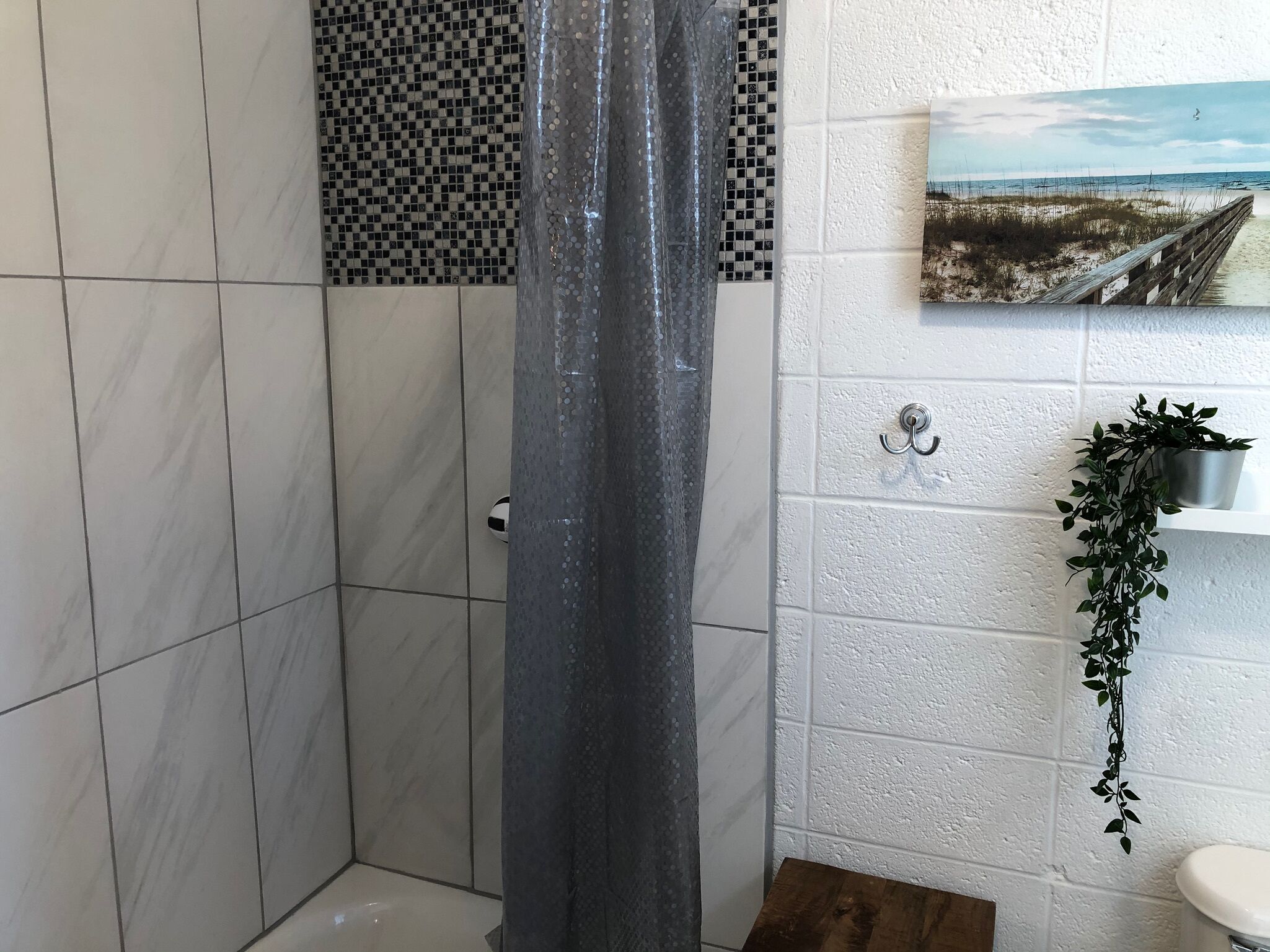 A bathroom with white tiles, a checkered tile backsplash, a shower curtain, a wooden bench, a hanging plant, and a picture on the wall.