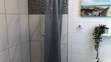 A bathroom with white tiles, a checkered tile backsplash, a shower curtain, a wooden bench, a hanging plant, and a picture on the wall.