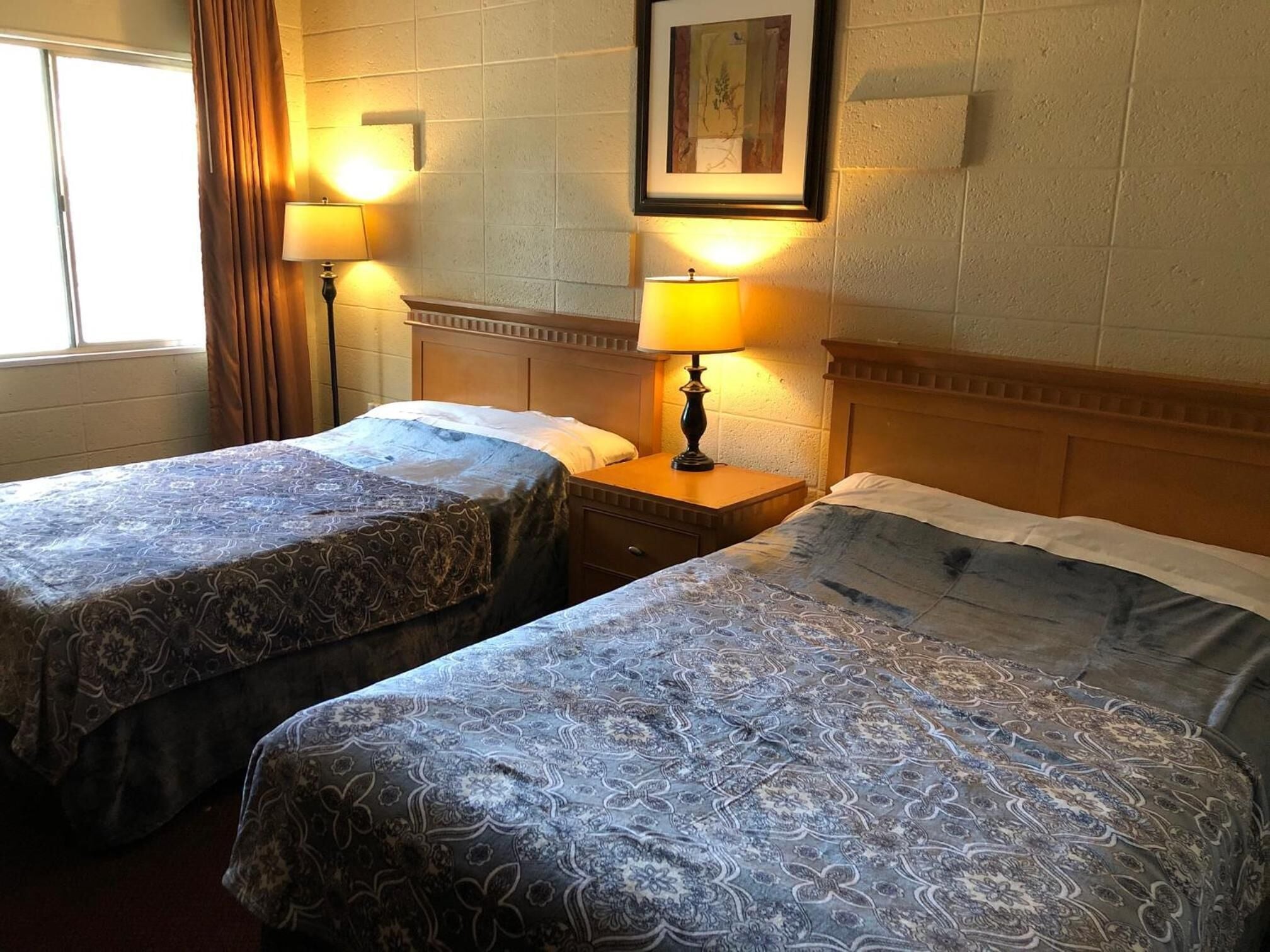 Quarto de hotel com duas camas de solteiro, cada uma com colcha estampada e mesa de cabeceira com abajur. Há uma moldura com um quadro na parede acima das camas.