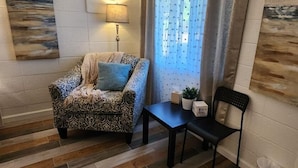 Uma sala de estar aconchegante com uma poltrona estampada, uma mesinha e uma cadeira preta. Há uma luminária sobre a mesa e um quadro na parede.