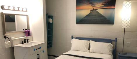 Habitación de hotel con cama, baño con tocador, un cuadro de un atardecer sobre el agua y una lámpara.