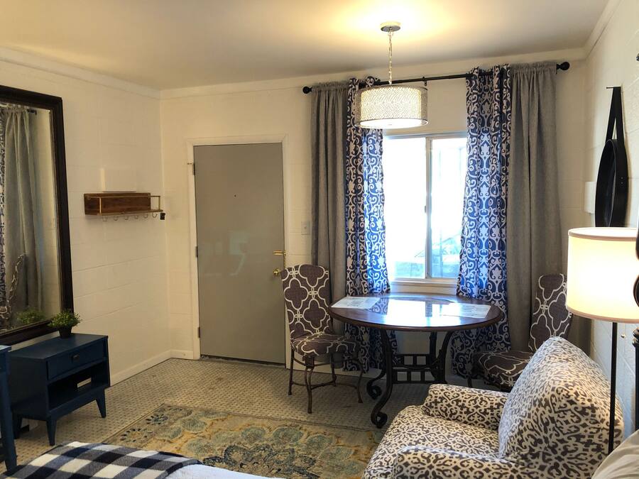A room with a round table, two patterned chairs, a sofa, a small side table, a door, and a window with curtains.