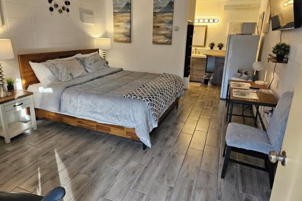 Une chambre avec un lit en bois, une table de chevet, une chaise et un bureau avec une lampe. On aperçoit une salle de bain au fond.