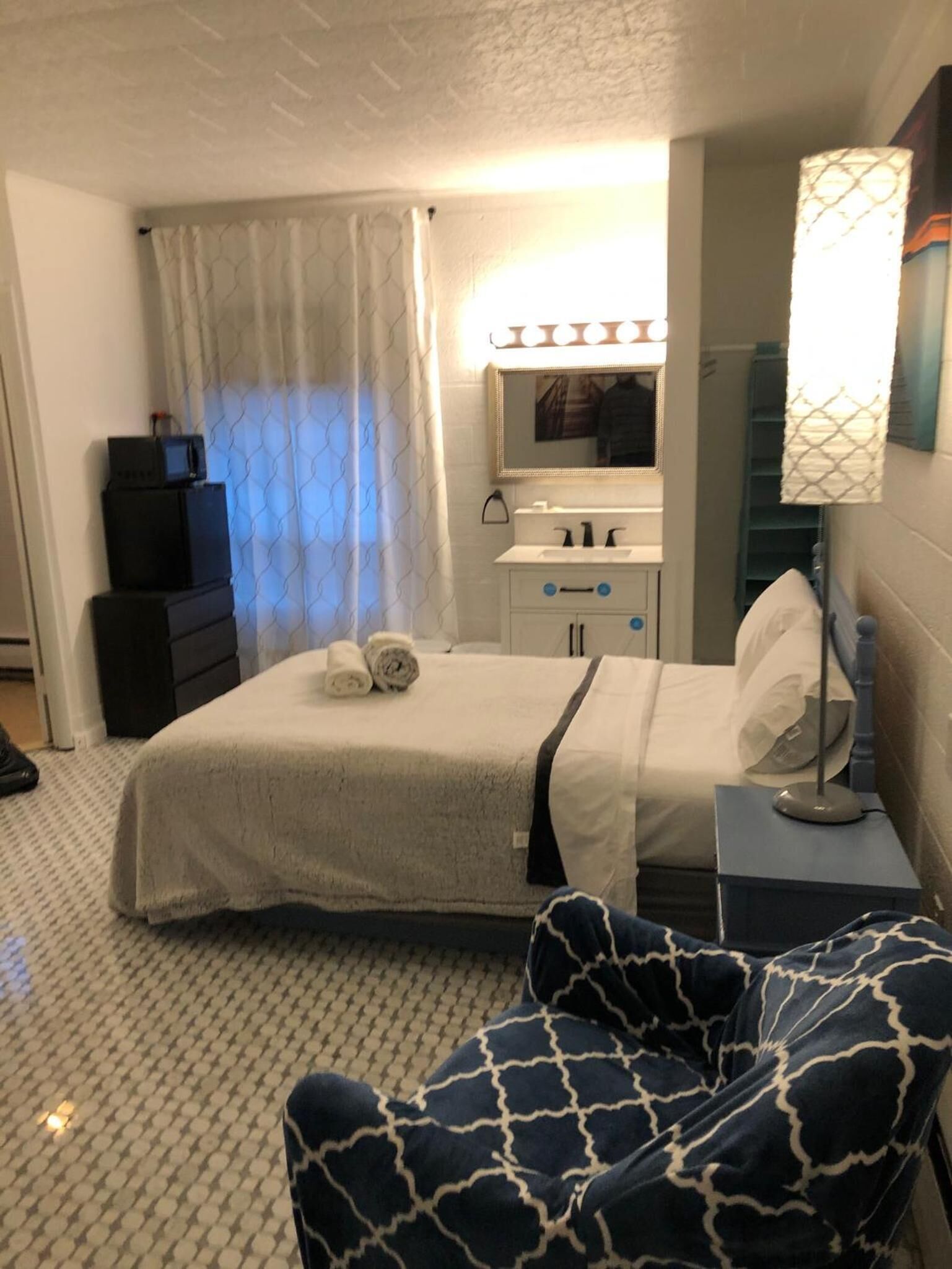 A hotel room with a bed, a desk, a chair, a mirror with lights, and a window with curtains.