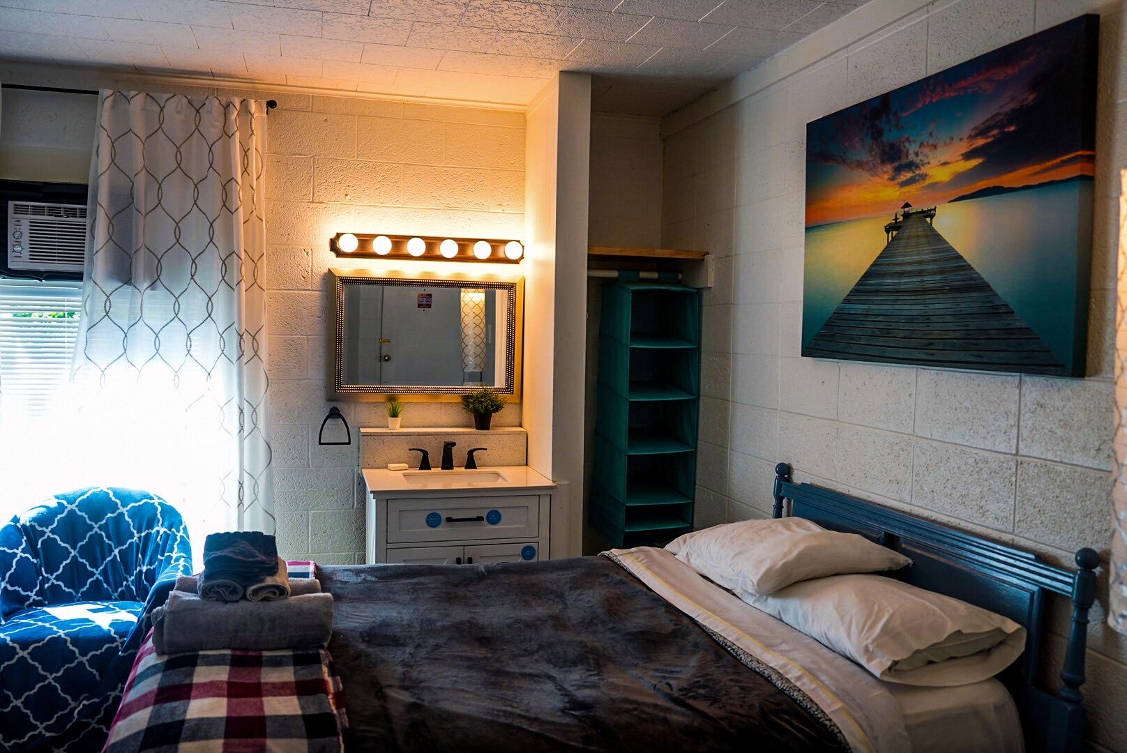 A bedroom with a bed, a vanity with a mirror, a shelf, and a painting of a pier.