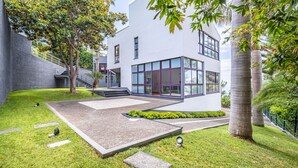 A modern building with a glass facade, surrounded by a well-maintained garden with grass, pathways, and trees. - Vila Crerital - By Wehost (Funchal)
